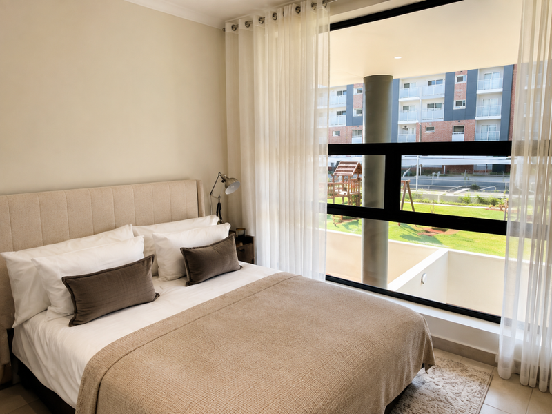 A well-lit bedroom featuring large floor-to-ceiling windows that provide ample natural light and views of the surrounding complex grounds.