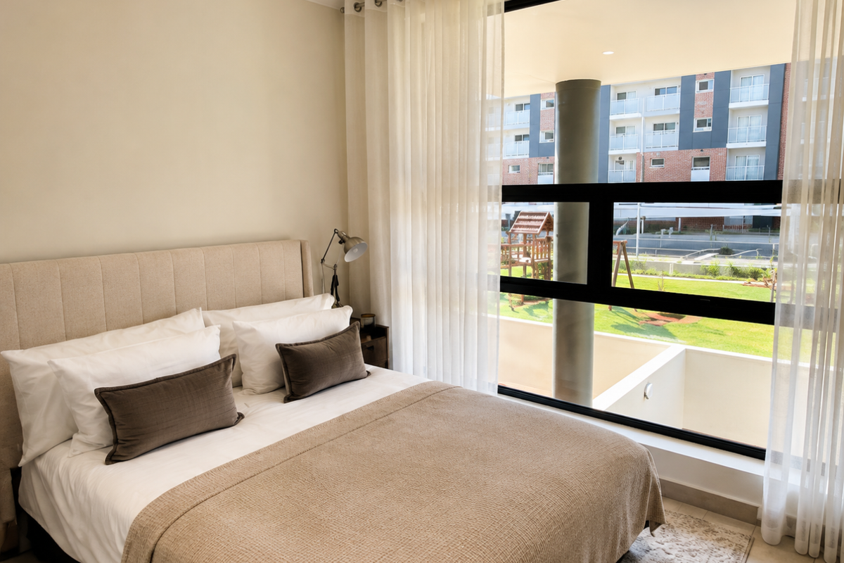 A well-lit bedroom featuring large floor-to-ceiling windows that provide ample natural light and views of the surrounding complex grounds.