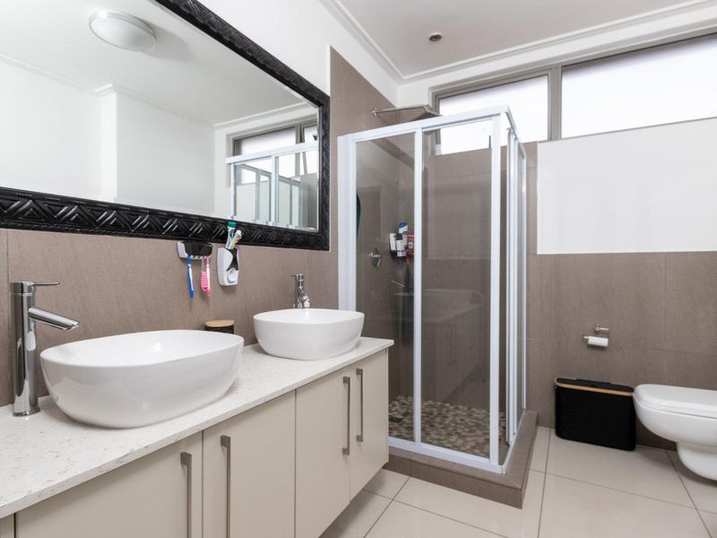 Modern bathroom featuring a double vanity with vessel sinks, a glass-enclosed shower, and a wall-mounted toilet with neutral tiled finishes.