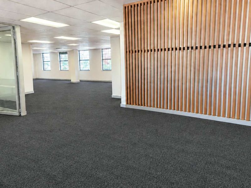 Spacious open-plan office area featuring a modern timber slat feature wall, glass partitioning, suspended ceilings with recessed lighting, and neutral carpet flooring.