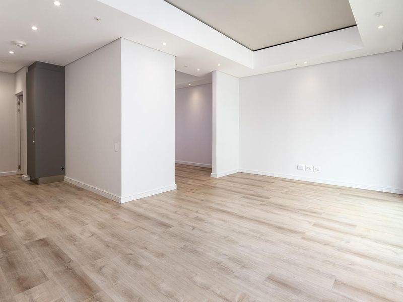 A bright, modern, and empty living area featuring light wood-look laminate flooring, recessed ceiling lighting, and clean white walls.