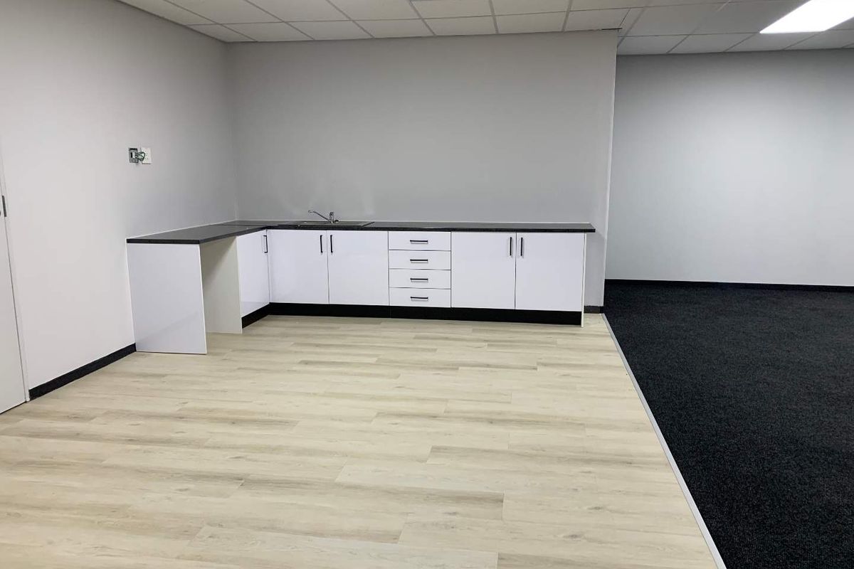 A clean, modern office kitchenette area featuring white cabinetry, dark countertops, a stainless steel sink, and a mix of wood-look vinyl and carpet flooring.