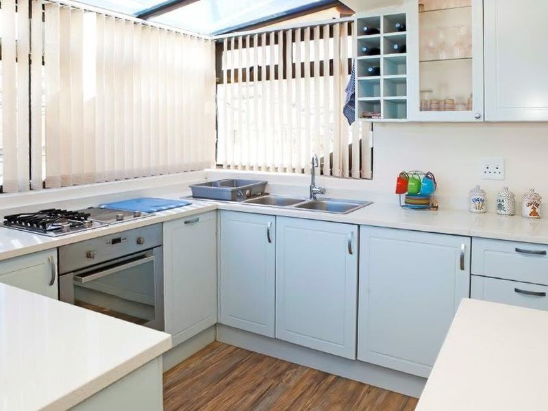 A bright kitchen featuring light blue cabinetry, a built-in oven, gas hob, and a stainless steel sink, complemented by wood-look flooring and vertical blinds.