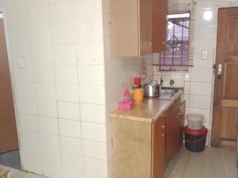 A compact kitchen area featuring tiled walls, wooden cabinetry, and a stainless steel sink, requiring cosmetic updates to modernize the space.