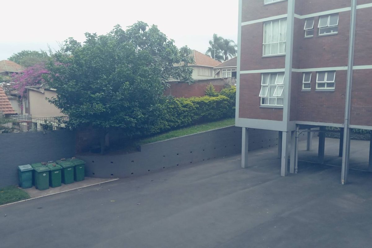 View of the apartment building exterior, showing the paved driveway, parking area, and surrounding landscaped retaining wall.