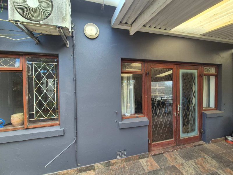 Covered patio area featuring tiled flooring, timber-framed doors and windows with security grilles, and an exterior-mounted air conditioning unit.