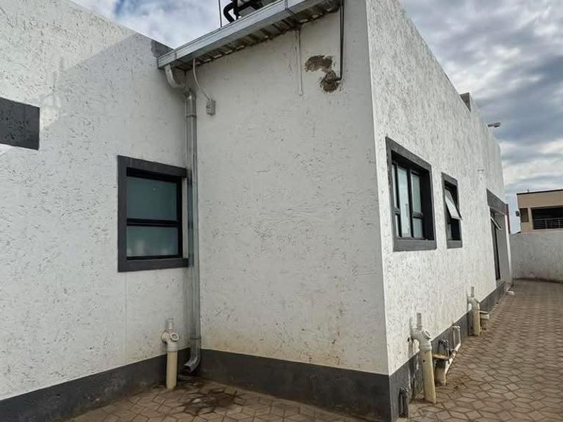 Rear exterior view of the property featuring a paved patio area, solar water heating system, and white textured wall finish with dark window frames.