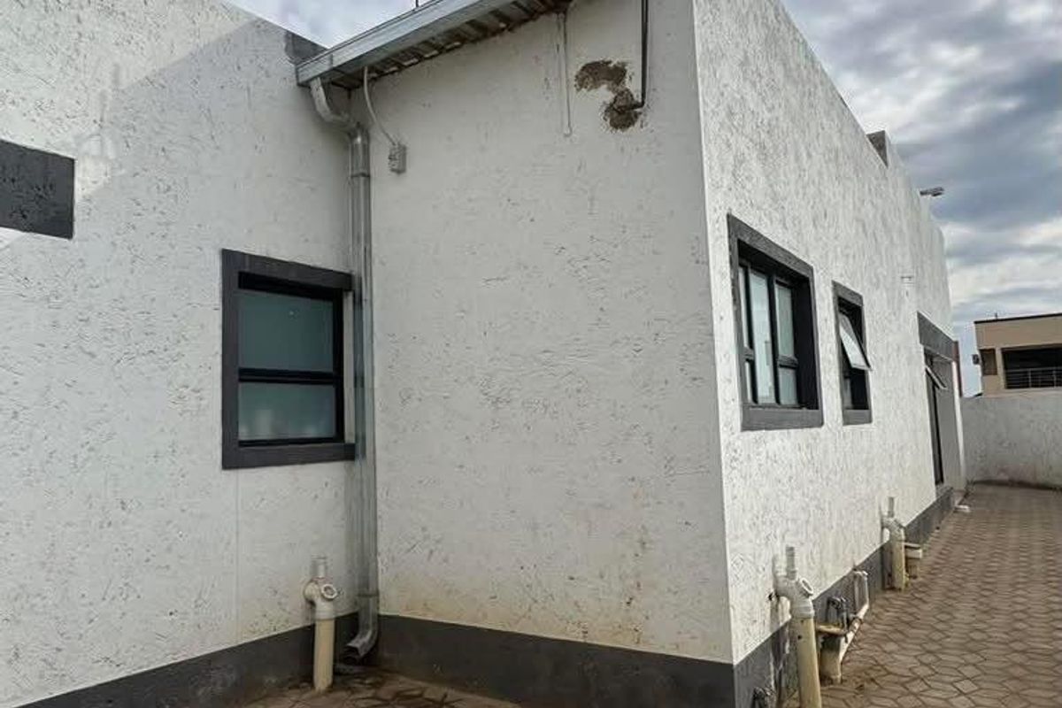 Rear exterior view of the property featuring a paved patio area, solar water heating system, and white textured wall finish with dark window frames.