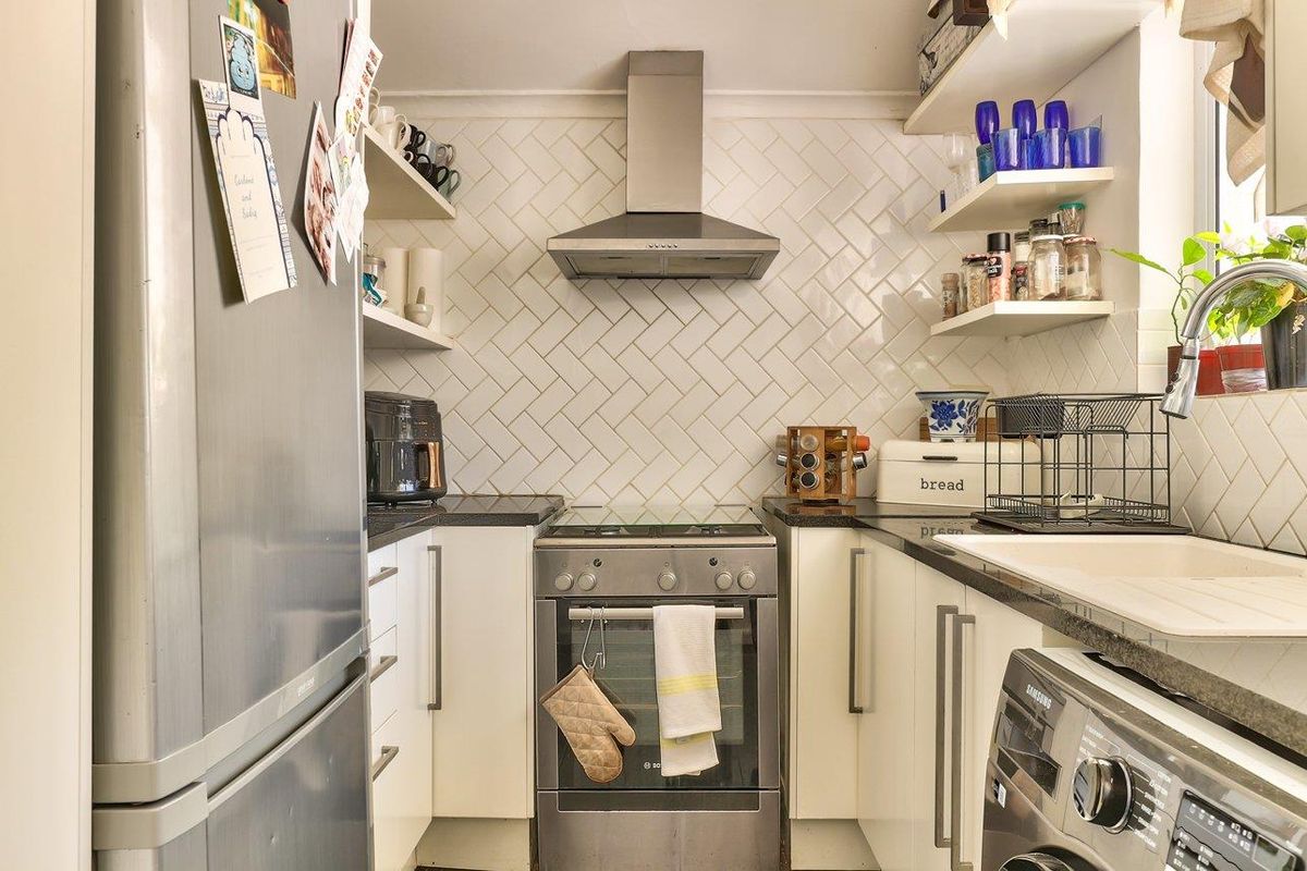 A compact, well-maintained kitchen featuring a stylish herringbone tile backsplash, stainless steel extractor fan, built-in oven, and efficient storage with floating shelves.