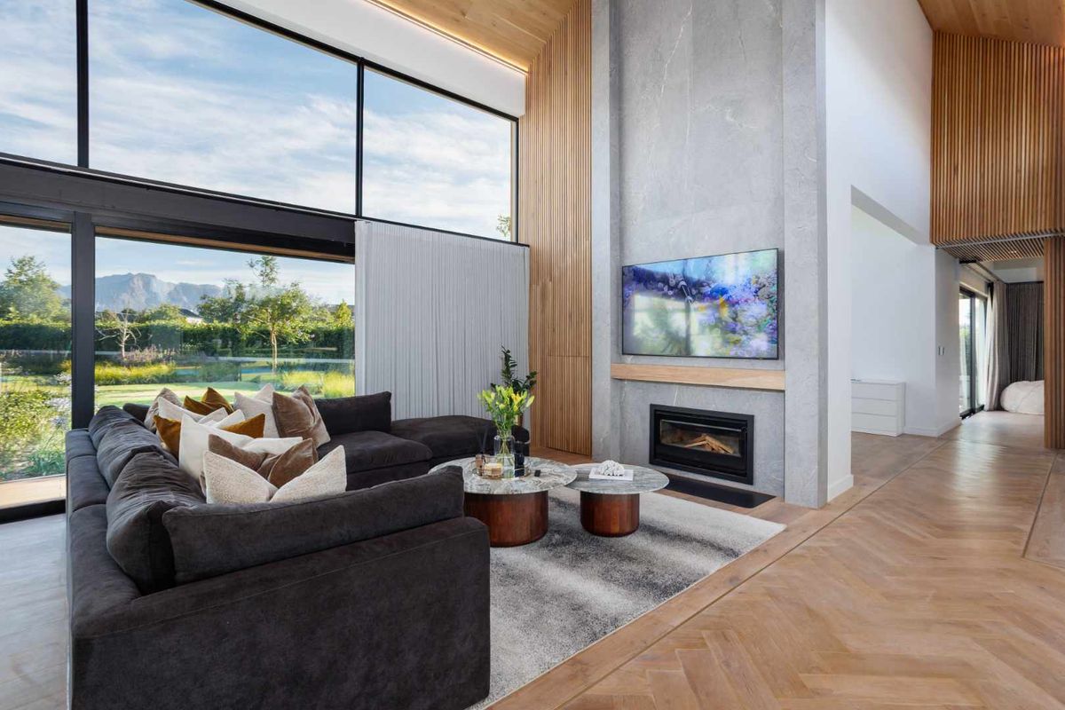 Spacious living room featuring high vaulted ceilings, herringbone timber flooring, a striking stone-clad fireplace, and expansive floor-to-ceiling windows with garden views.