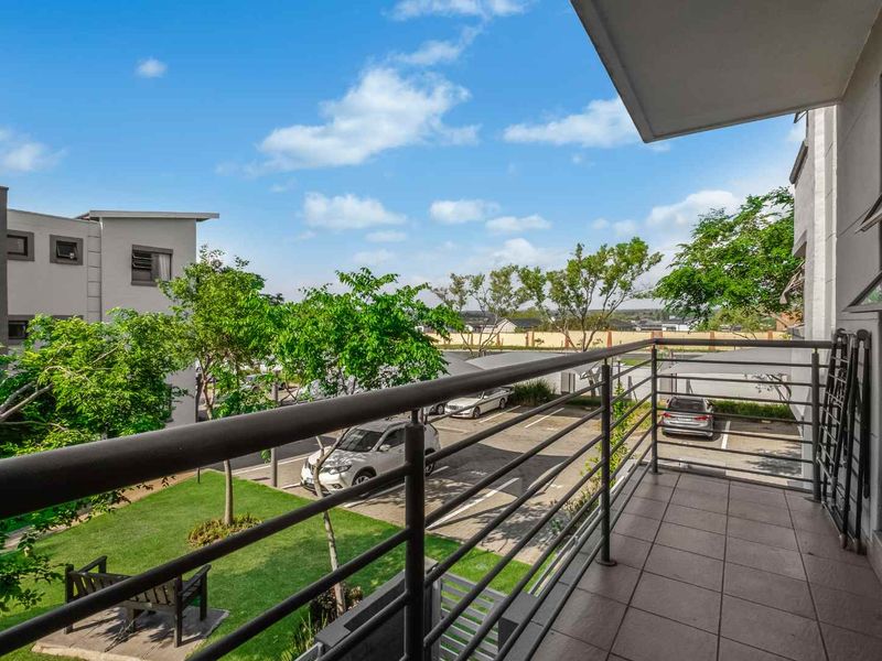 A clean, tiled balcony featuring a metal balustrade, offering an elevated view of the apartment complex grounds and parking area.