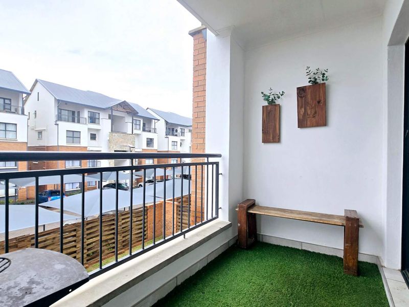 A covered balcony featuring a metal balustrade, artificial grass flooring, and direct access to the apartment interior via a sliding door.