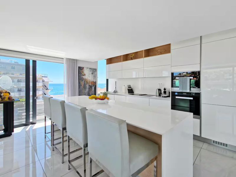 Modern open-plan kitchen featuring a sleek island, high-gloss cabinetry, integrated appliances, and large sliding doors opening to a scenic ocean view.