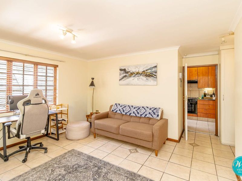 A living room area with tiled flooring and natural light, featuring an adjacent kitchen with wooden cabinetry visible through the doorway.