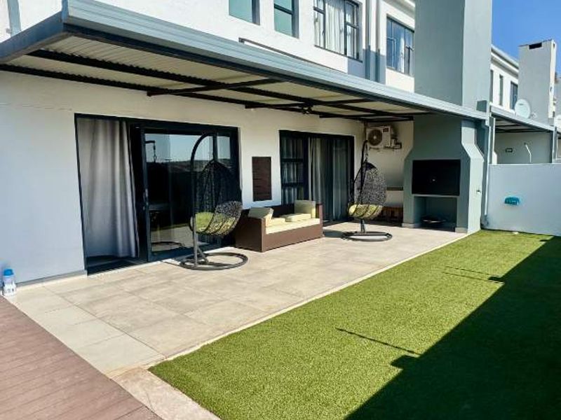Rear exterior view of the townhouse featuring a covered patio with a built-in braai, artificial lawn, and timber decking.