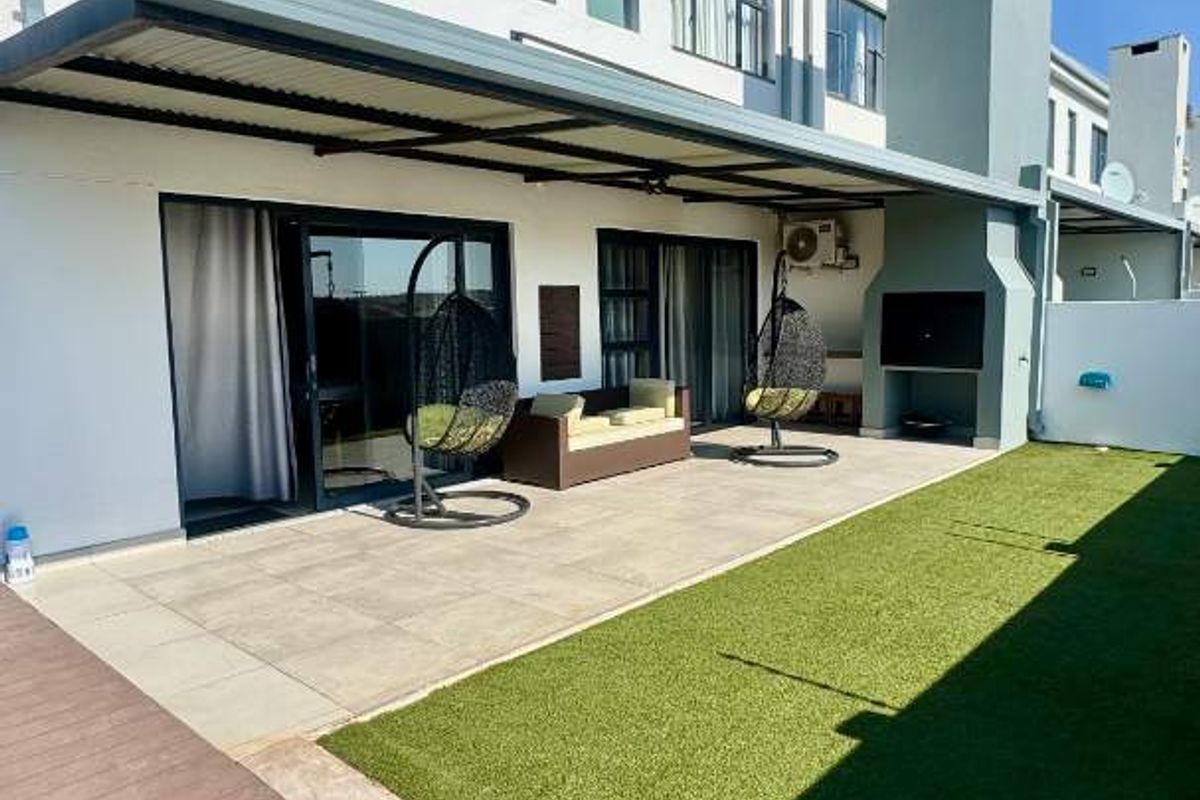 Rear exterior view of the townhouse featuring a covered patio with a built-in braai, artificial lawn, and timber decking.