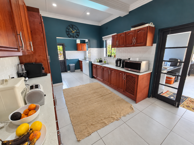 A spacious kitchen featuring dark wood cabinetry, white subway tile backsplash, stone countertops, and a connected scullery area with tiled flooring throughout.