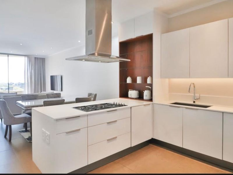 Modern open-plan kitchen and living area featuring sleek white cabinetry, a gas hob with extractor, and integrated sink, flowing into a bright dining space.
