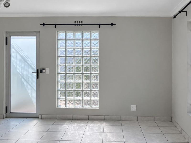 A bright, empty living area featuring neutral tiled flooring, a decorative glass block window, and secure access points with security bars and a frosted door.