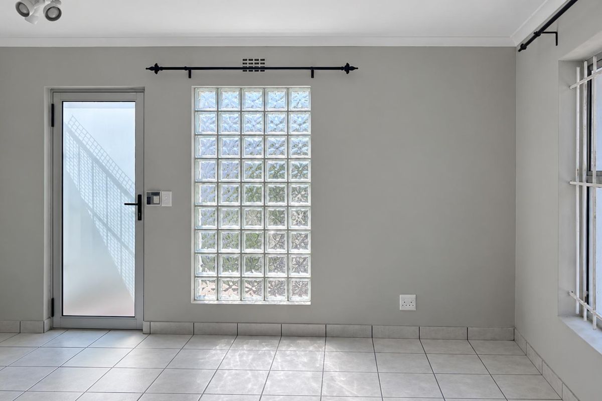 A bright, empty living area featuring neutral tiled flooring, a decorative glass block window, and secure access points with security bars and a frosted door.