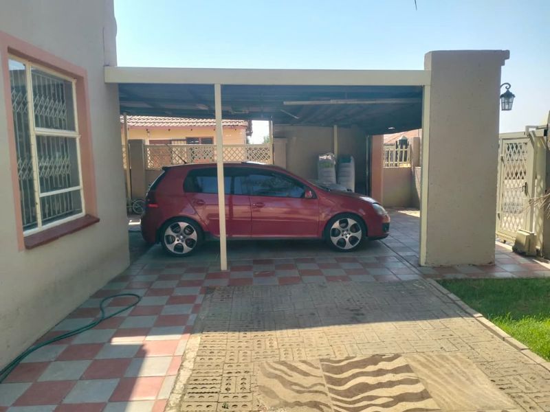 A covered carport area with tiled flooring and paved driveway, featuring security gates and bars on the adjacent building structure.