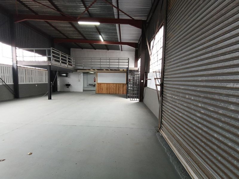 Spacious industrial warehouse unit featuring a large roller shutter door, mezzanine level with steel railing, and concrete flooring suitable for various commercial operations.