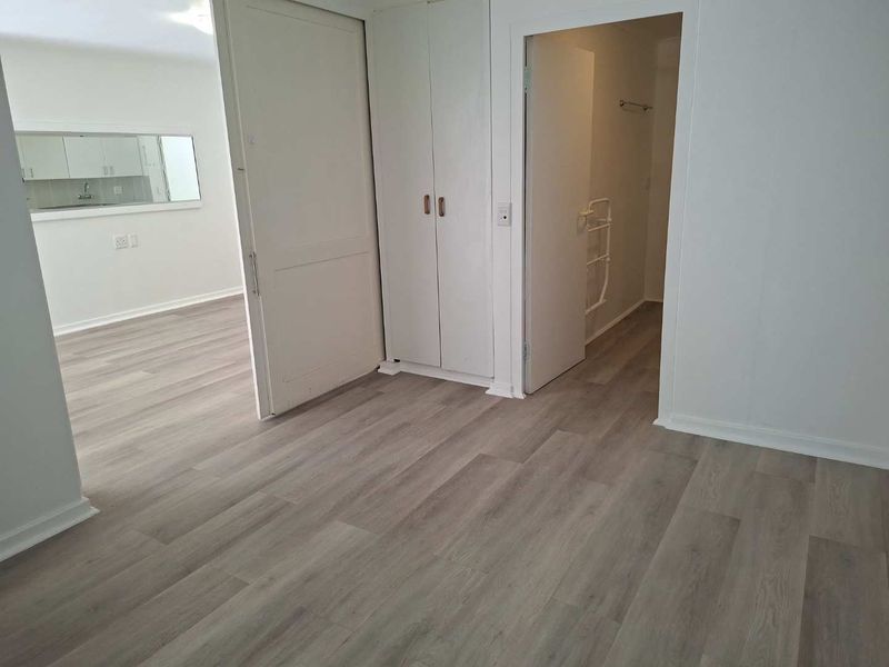 A bright, empty bedroom featuring modern light-toned laminate flooring and built-in cupboards, with an adjacent view into a kitchen and bathroom area.