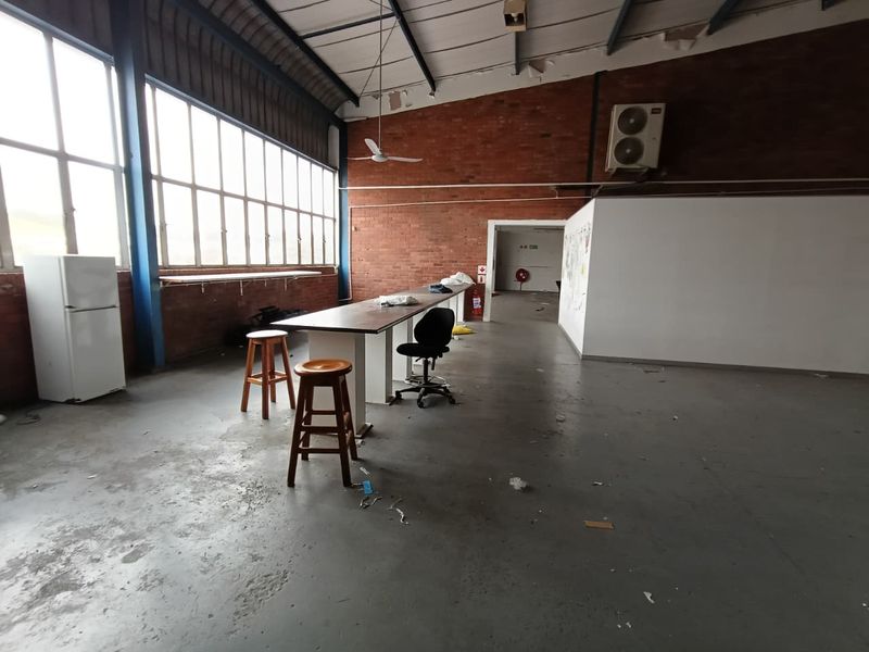 Spacious industrial open-plan area featuring exposed brick walls, high ceilings with steel trusses, large windows, and concrete flooring.