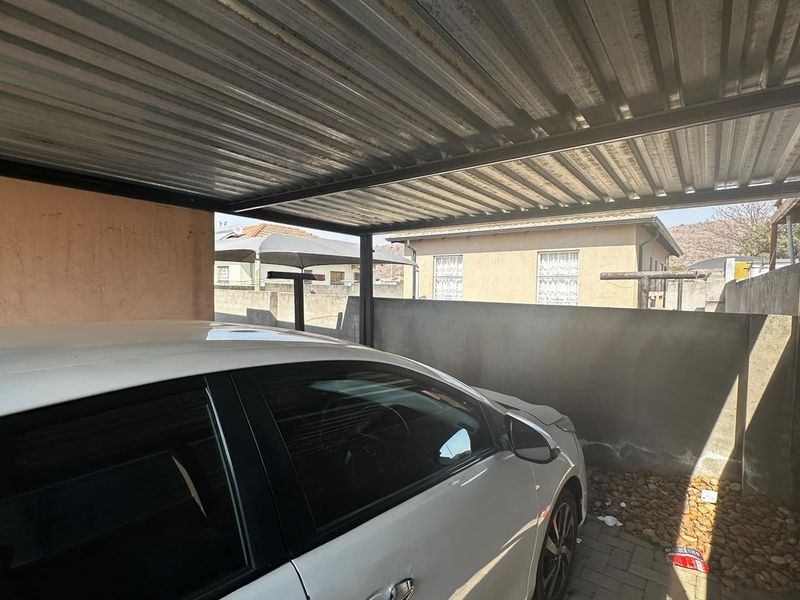 A functional outdoor carport featuring a corrugated metal roof supported by a steel frame, situated within a paved, walled perimeter area.