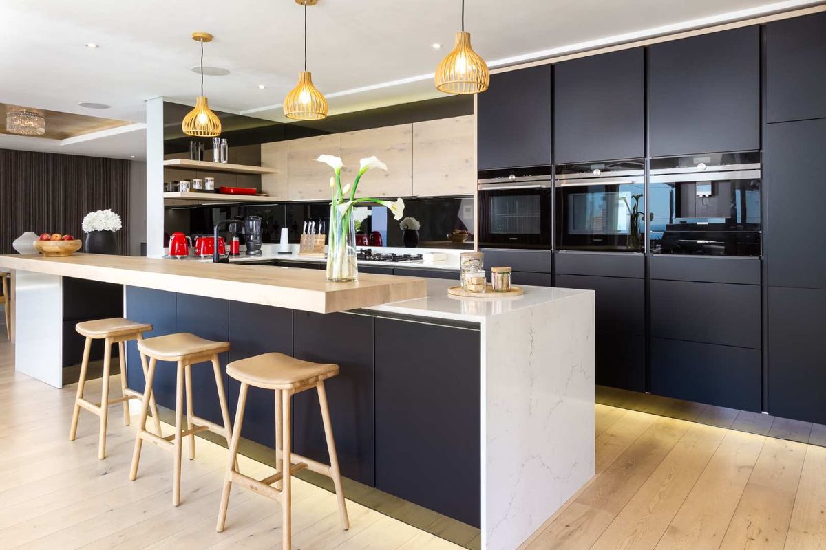 Modern kitchen featuring sleek matte black cabinetry, a stone island with a timber breakfast bar, integrated ovens, and high-quality timber flooring.