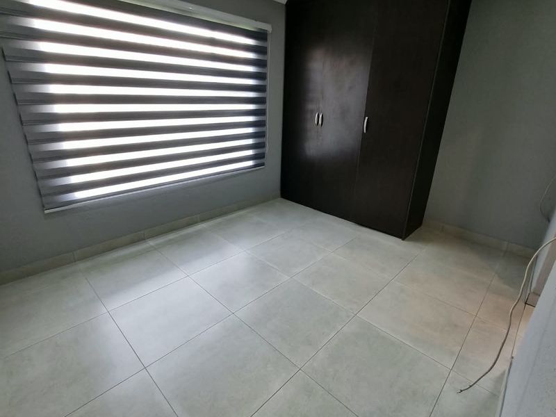 A clean, empty bedroom featuring neutral tiled flooring, recessed downlighting, modern zebra blinds, and a dark wood built-in wardrobe.