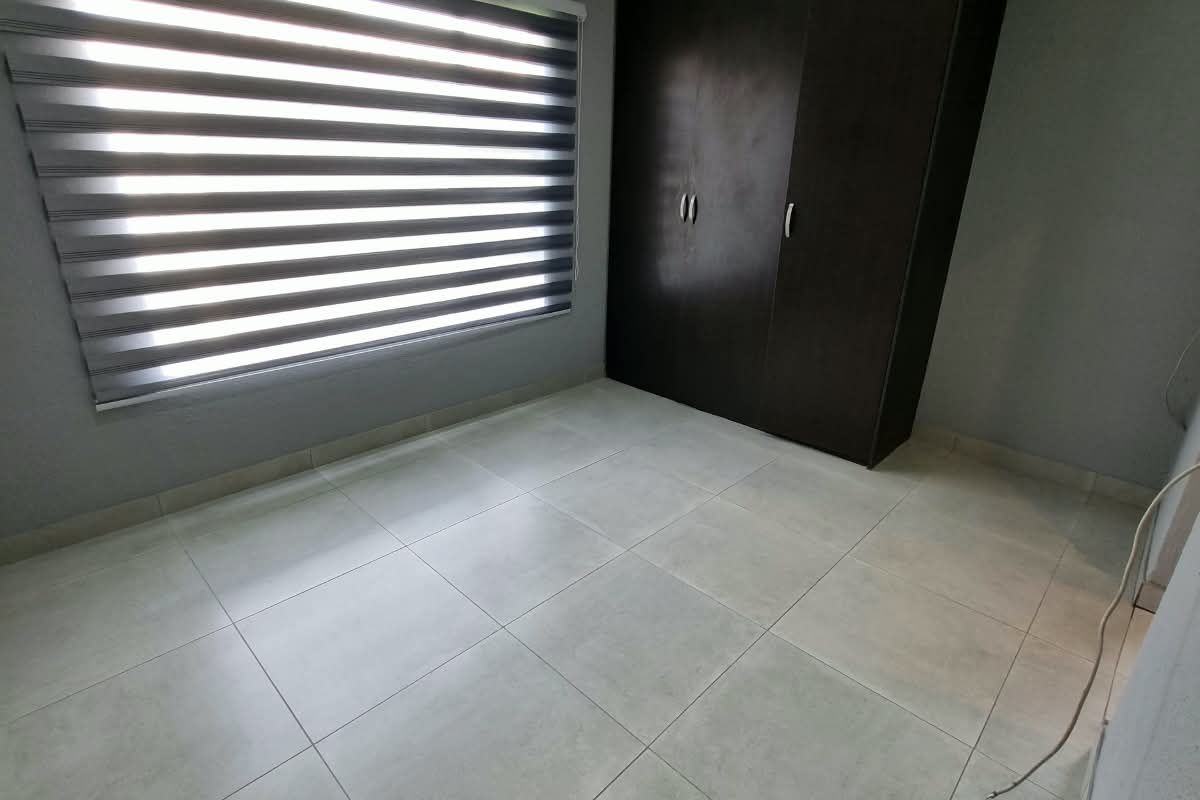 A clean, empty bedroom featuring neutral tiled flooring, recessed downlighting, modern zebra blinds, and a dark wood built-in wardrobe.