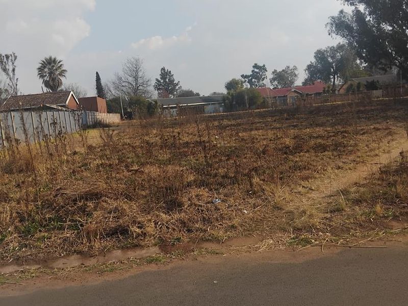 A vacant residential plot of land with road frontage, featuring dry, overgrown vegetation and surrounding residential neighborhood views.