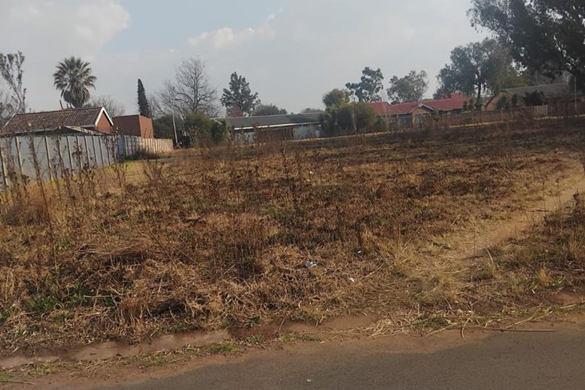 A vacant residential plot of land with road frontage, featuring dry, overgrown vegetation and surrounding residential neighborhood views.