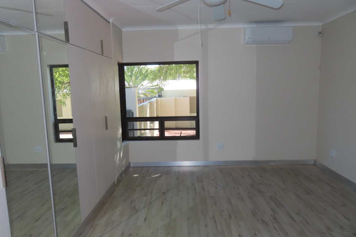 A bright bedroom featuring built-in mirrored wardrobes, laminate flooring, a ceiling fan, and a wall-mounted air conditioning unit.