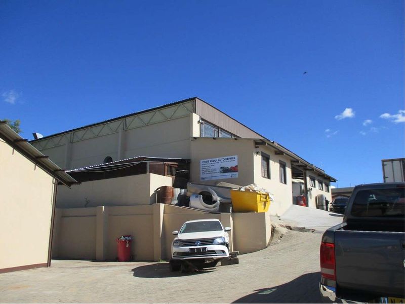 Exterior view of an industrial property featuring a concrete driveway, metal roof structure, and perimeter boundary wall.
