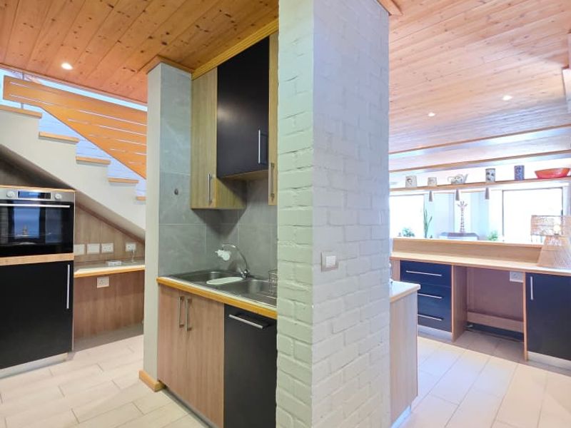 Modern kitchen featuring fitted cabinetry, built-in oven, stainless steel sink, and a distinctive exposed brick pillar under a timber-clad ceiling.
