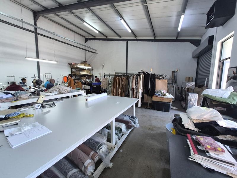 Industrial workspace featuring high ceilings with exposed steel trusses, concrete flooring, and functional fluorescent lighting, currently utilized as a garment production area.