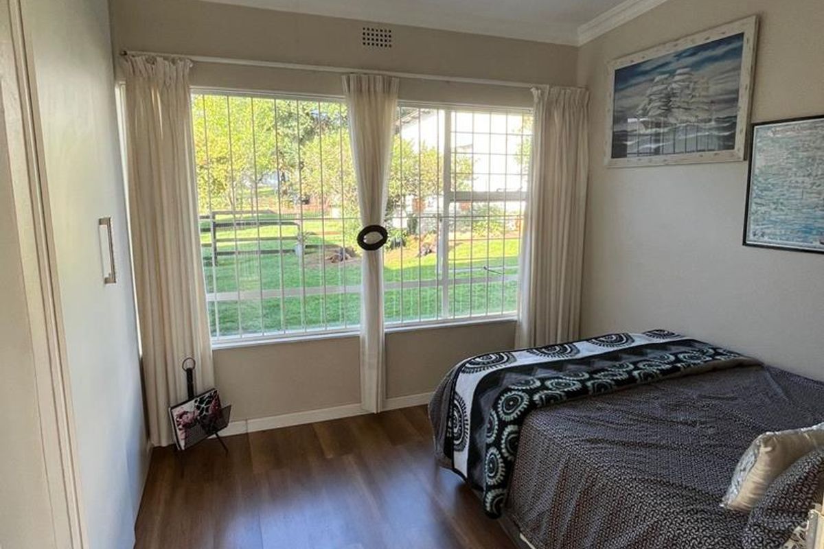 A well-lit bedroom featuring wood-look flooring, built-in cupboards, and large windows with security bars overlooking the garden.