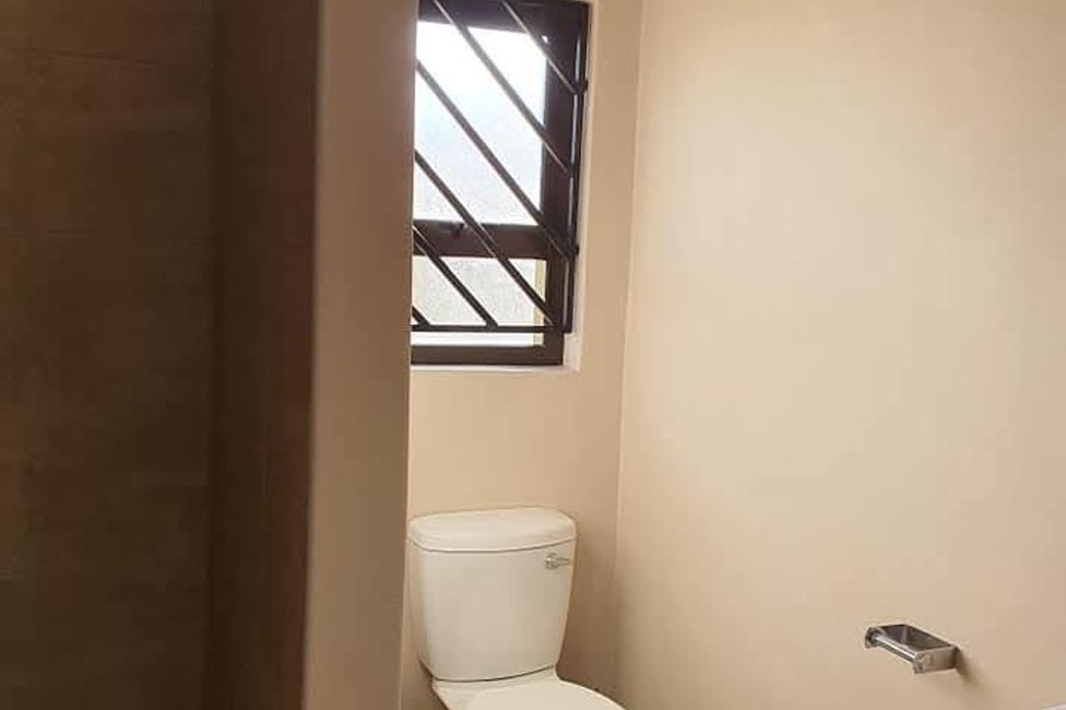 A functional bathroom featuring a tiled shower area, a standard toilet, and a window with security bars.