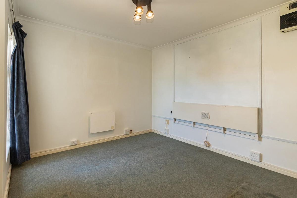 An empty bedroom featuring grey carpeting, wall-mounted heating and electrical fixtures, and a ceiling-mounted light fitting.