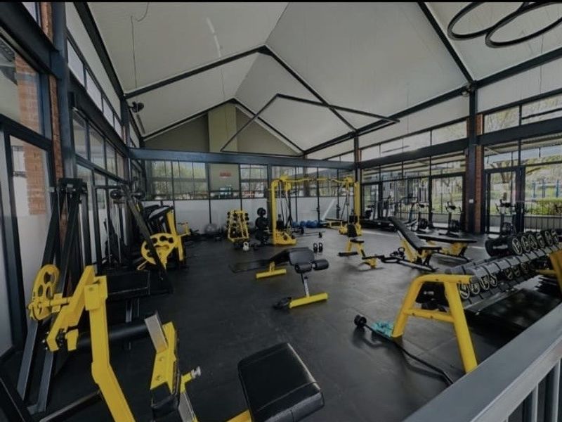A spacious, well-lit gym facility featuring high vaulted ceilings, exposed steel structural elements, and extensive floor-to-ceiling windows.