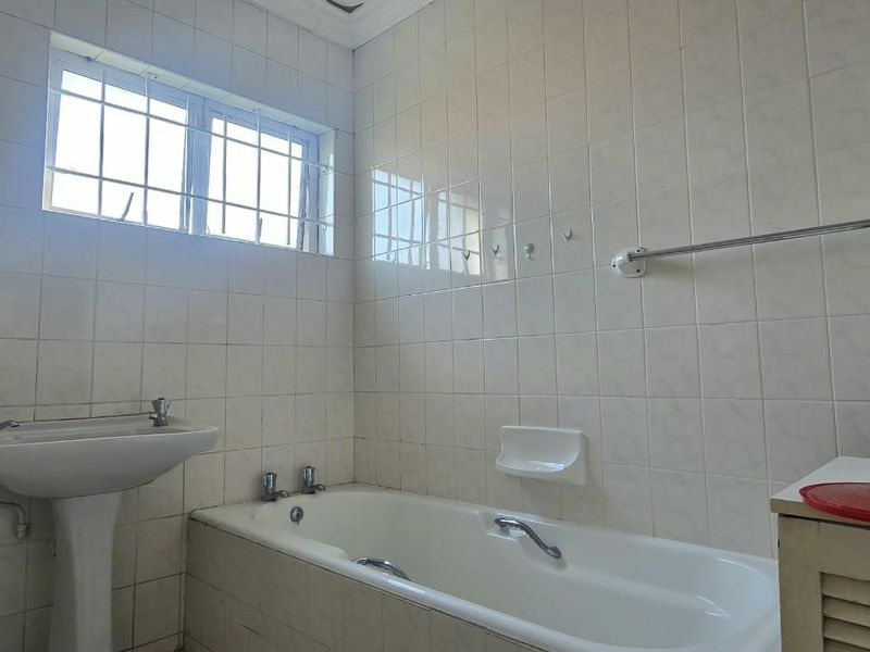 A dated bathroom featuring a pedestal basin, built-in bathtub, and full-height wall tiling, requiring cosmetic updates to modernize the space.