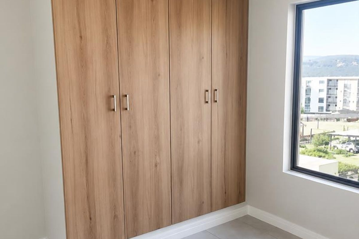 A bright, modern bedroom featuring neutral tiled flooring, built-in wood-finish cupboards, and a large window offering natural light and exterior views.