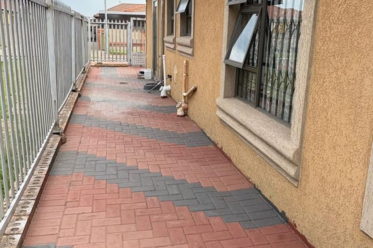 A side exterior walkway featuring patterned brick paving, security fencing, and windows with fitted security bars.