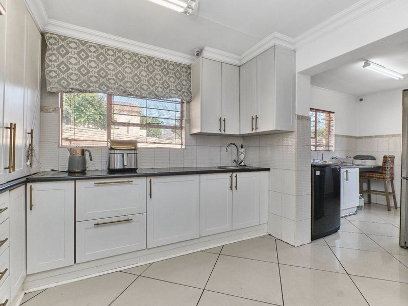 A functional kitchen featuring light-toned cabinetry, dark countertops, tiled backsplash, and ceramic flooring, with an adjacent scullery area visible.