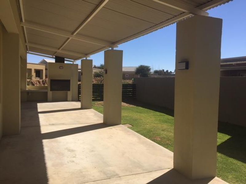 A spacious covered patio featuring a built-in braai, concrete flooring, and support pillars, overlooking a private garden area.
