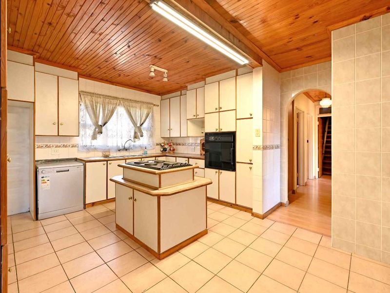 A spacious, dated kitchen featuring timber ceilings, tiled flooring, a central island with a cooktop, and built-in cabinetry with an integrated oven.