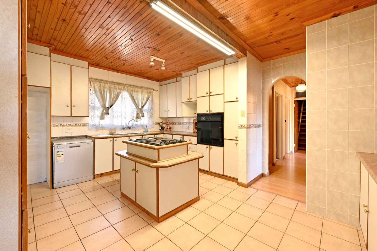 A spacious, dated kitchen featuring timber ceilings, tiled flooring, a central island with a cooktop, and built-in cabinetry with an integrated oven.