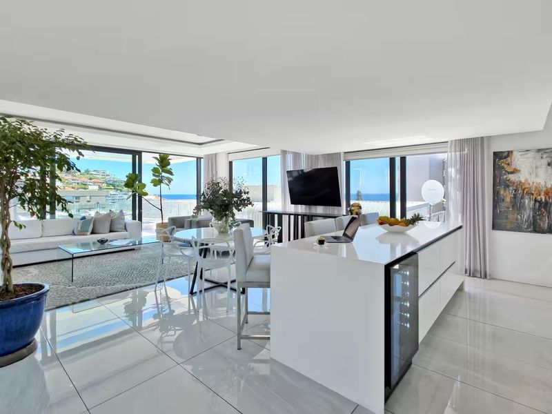 Spacious open-plan living and kitchen area featuring a modern island, sleek tiled floors, and expansive floor-to-ceiling glass doors with panoramic coastal views.